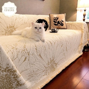 May include: Cream-coloured sofa cover with a floral pattern. A white cat is lying on the sofa. The cover has a textured appearance and a decorative border. A pillow with a floral design and a cup of coffee are also visible.