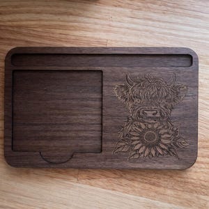 Puede incluir: Una bandeja rectangular de madera con un dise&ntilde;o tallado de una vaca de las Tierras Altas y un girasol. La bandeja tiene un &aacute;rea empotrada y una peque&ntilde;a ranura. La madera es de color marr&oacute;n oscuro.