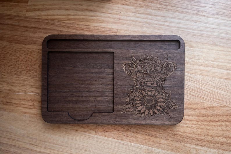 Puede incluir: Una bandeja rectangular de madera con un dise&ntilde;o tallado de una vaca de las Tierras Altas y un girasol. La bandeja tiene un &aacute;rea empotrada y una peque&ntilde;a ranura. La madera es de color marr&oacute;n oscuro.