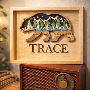 Puede incluir: Arte de pared de madera con la silueta de un oso y una escena de montaña y bosque. El oso está tallado con un acabado de madera natural, y las montañas y los árboles están pintados en verde, azul y negro. La palabra "TRACE" está tallada debajo.