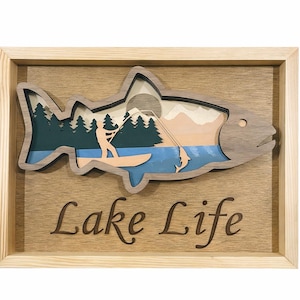 Puede incluir: Arte de pared enmarcado en madera con un diseño en forma de pez que representa una escena de lago. La obra de arte incluye una persona practicando paddle surf, árboles, montañas y las palabras "Lake Life". El marco es marrón claro.