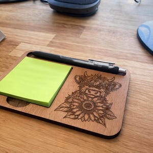Puede incluir: Un organizador de escritorio rectangular de madera con una pila de notas adhesivas de color verde ne&oacute;n a la izquierda y un grabado l&aacute;ser de una vaca Highland con un girasol a la derecha, con un bol&iacute;grafo negro en la ranura superior.