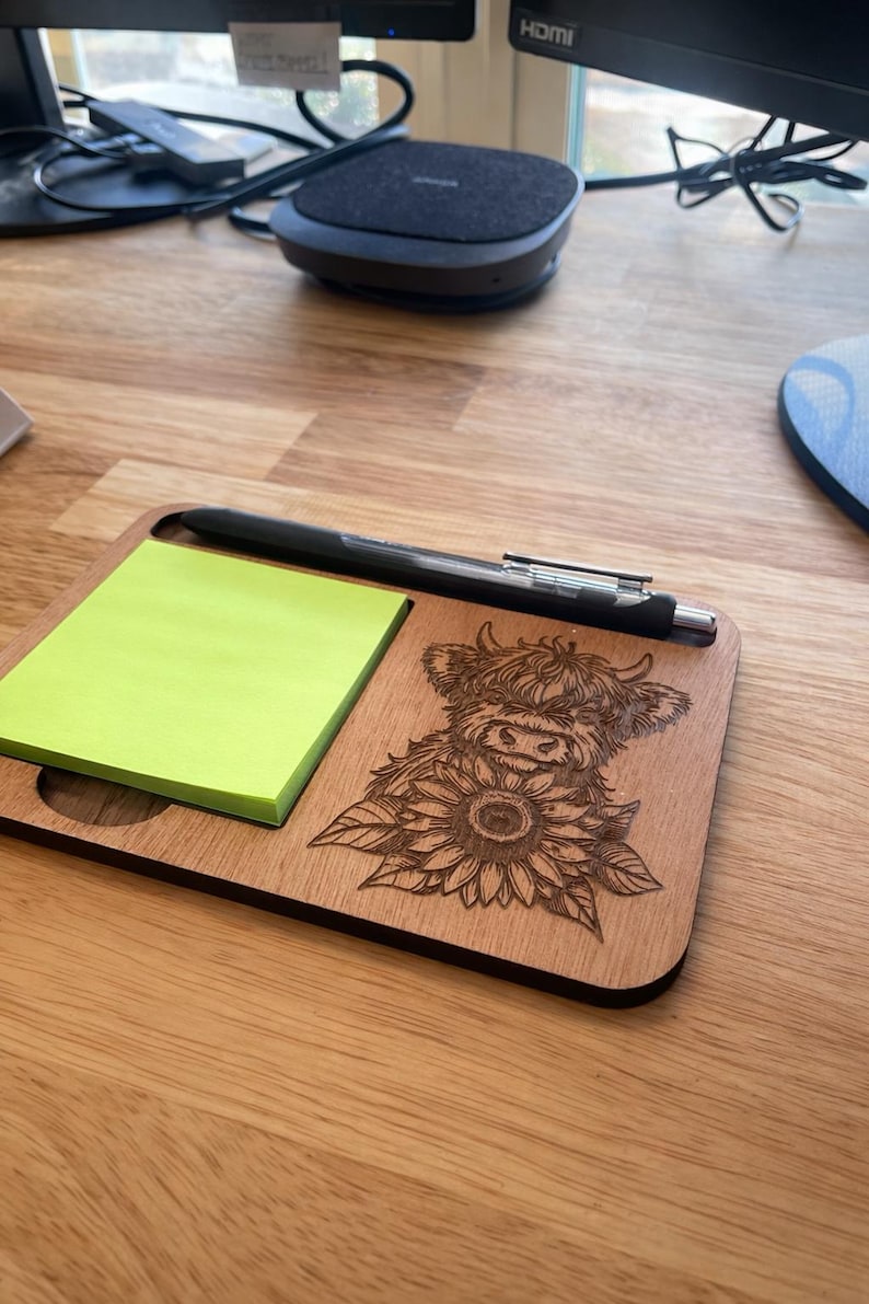 Puede incluir: Un organizador de escritorio rectangular de madera con una pila de notas adhesivas de color verde ne&oacute;n a la izquierda y un grabado l&aacute;ser de una vaca Highland con un girasol a la derecha, con un bol&iacute;grafo negro en la ranura superior.