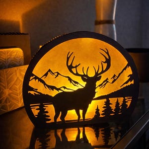 Puede incluir: Una lámpara decorativa circular negra con la silueta de un ciervo frente a un paisaje de montañas y bosque. La lámpara emite un brillo amarillo cálido, creando un ambiente acogedor. La lámpara está colocada sobre una superficie reflectante.