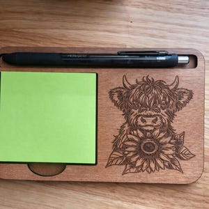 Puede incluir: Organizador de escritorio de madera con ranura para bolígrafo, bloc de notas adhesivas verde y diseño grabado con láser de una vaca de las Tierras Altas con un girasol. Un bolígrafo negro descansa en la ranura superior. El organizador es de madera marrón.