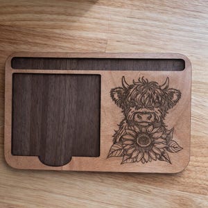 Puede incluir: Un organizador de escritorio rectangular de madera con una bandeja empotrada de color marr&oacute;n oscuro a la izquierda y un panel de madera m&aacute;s claro a la derecha. La parte derecha muestra un grabado l&aacute;ser de una vaca Highland y un girasol.