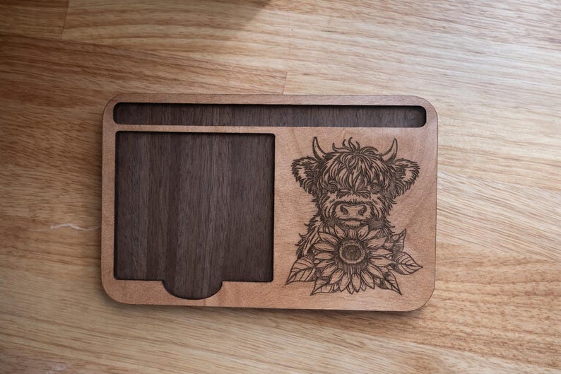 Puede incluir: Un organizador de escritorio rectangular de madera con una bandeja empotrada de color marr&oacute;n oscuro a la izquierda y un panel de madera m&aacute;s claro a la derecha. La parte derecha muestra un grabado l&aacute;ser de una vaca Highland y un girasol.