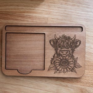 Puede incluir: Bandeja de madera con un dise&ntilde;o tallado de una vaca de las Tierras Altas y un girasol. La bandeja tiene forma rectangular con un &aacute;rea empotrada y un recorte curvo. La madera tiene un tono c&aacute;lido y natural.