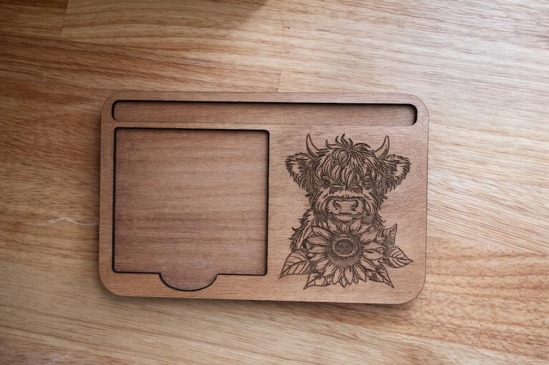 Puede incluir: Bandeja de madera con un dise&ntilde;o tallado de una vaca de las Tierras Altas y un girasol. La bandeja tiene forma rectangular con un &aacute;rea empotrada y un recorte curvo. La madera tiene un tono c&aacute;lido y natural.