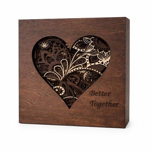 Puede incluir: Una caja de madera marrón oscuro con un recorte en forma de corazón que revela un intrincado diseño floral. El texto "Better Together" está grabado en la parte delantera. El diseño presenta patrones en capas y una veta de madera natural.