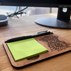 Puede incluir: Organizador de escritorio de madera con un dise&ntilde;o grabado con l&aacute;ser de una vaca de las Tierras Altas y girasoles. El organizador contiene un bloc de notas adhesivas amarillas y un bol&iacute;grafo negro. La madera tiene un acabado natural.