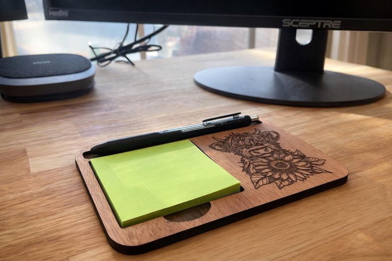 Puede incluir: Organizador de escritorio de madera con un dise&ntilde;o grabado con l&aacute;ser de una vaca de las Tierras Altas y girasoles. El organizador contiene un bloc de notas adhesivas amarillas y un bol&iacute;grafo negro. La madera tiene un acabado natural.