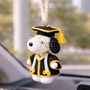 Puede incluir: Un adorno de coche de Snoopy tejido a ganchillo, vestido con una toga y birrete de graduación. La toga negra tiene ribetes amarillos y detalles en azul claro. El birrete tiene una borla amarilla. El adorno cuelga de un cordón trenzado color crema.