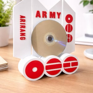 May include: A white and red decorative CD holder with the word "ARMY" and "ARIRANG" in red letters. A gold CD is inserted in the holder. The holder is on a wooden surface.
