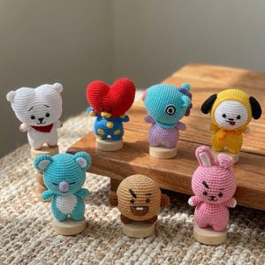 May include: A collection of seven colorful, crocheted character figures. Each figure is a different color and design, including a white bear, a red heart, a blue figure with a purple headpiece, a yellow figure with black ears, a blue koala, a brown figure, and a pink bunny.
