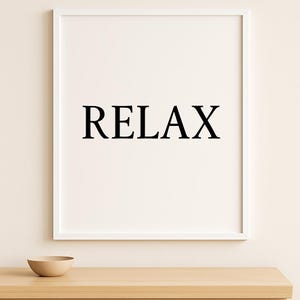 Puede incluir: Un cuadro enmarcado con la palabra "RELAX" en letras negras en negrita centrada sobre un fondo blanco. El marco es blanco, y un peque&ntilde;o cuenco beige descansa sobre una superficie de madera debajo. Tema: decoraci&oacute;n del hogar.