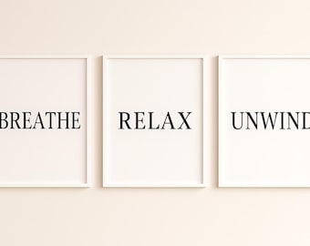 May include: Three framed art prints with the words "Breathe", "Relax", and "Unwind" in a black, sans-serif font. The prints have white backgrounds and white frames, creating a minimalist aesthetic. The artwork is displayed against a neutral-colored wall.