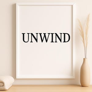 Puede incluir: Una impresi&oacute;n enmarcada con la palabra "UNWIND" en letras negras en negrita. La impresi&oacute;n est&aacute; en un marco blanco y se muestra sobre una superficie de madera clara. Un jarr&oacute;n beige con plantas secas y una manta doblada tambi&eacute;n est&aacute;n en la superficie.