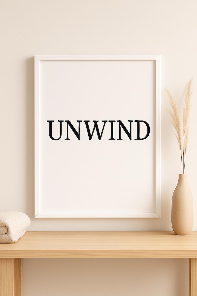 Puede incluir: Una impresi&oacute;n enmarcada con la palabra "UNWIND" en letras negras en negrita. La impresi&oacute;n est&aacute; en un marco blanco y se muestra sobre una superficie de madera clara. Un jarr&oacute;n beige con plantas secas y una manta doblada tambi&eacute;n est&aacute;n en la superficie.