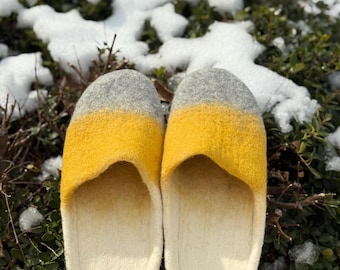 May include: A pair of felt slippers with a color block design. The slippers have a white sole, a bright yellow midsection, and a gray top. The slippers are resting on a bed of green foliage with patches of snow.