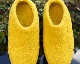 May include: A pair of bright yellow felt slippers. The slippers have a rounded toe and a simple, open design. The felt material appears soft and textured. The background is blurred, with green foliage and a reflective surface.