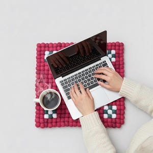 May include: A laptop sits on a red felted wool square with green and white accents. A white mug of coffee steams beside the laptop. The image is taken from an overhead perspective.
