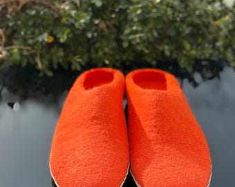 May include: A pair of bright orange felt slippers. The slippers have a rounded toe and a low profile. The soles are a light beige color. The background features green foliage and snow.