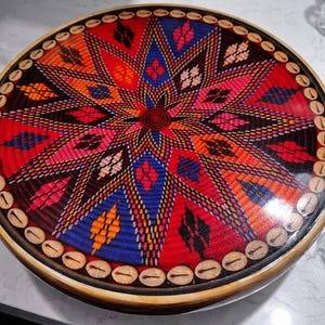 May include: A round, woven tray with a vibrant, starburst design in red, blue, orange, and pink. The tray's edge is adorned with small, light-colored shell accents. The base is a light brown color. This decorative tray is perfect for serving or display.
