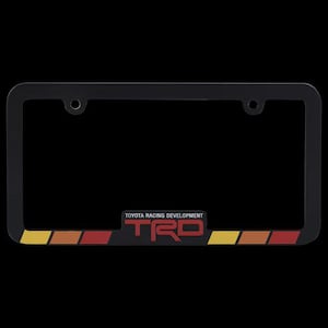 Puede incluir: Marco de matrícula negro con el texto "TOYOTA RACING DEVELOPMENT" y "TRD" en rojo. El marco tiene rayas amarillas, naranjas y rojas en la parte inferior.