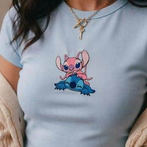 May include: Pale blue t-shirt with an embroidered design of two cartoon characters, one pink and one blue. The pink character is on top of the blue character. A gold necklace with a key pendant is visible.