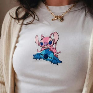 May include: White t-shirt with an embroidered design of two cartoon characters, one pink and one blue. The pink character is on top of the blue character. The shirt has a crew neck.
