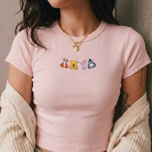 May include: A light pink t-shirt with a crew neck and short sleeves. The shirt features a graphic of four cartoon characters hanging on a string. The characters are a tiger, a bear, a pig, and a donkey.