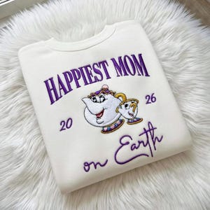 May include: Cream sweatshirt with purple embroidered text reading "Happiest Mom on Earth". The design includes a cartoon teapot and teacup. The numbers "20" and "26" are also embroidered on the sweatshirt.