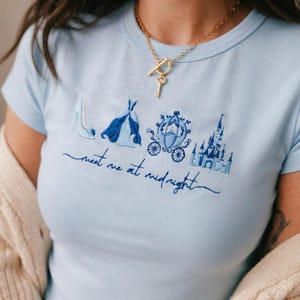 May include: Light blue t-shirt with embroidered Cinderella-themed design. Features a high heel, gown, carriage, and castle. The text "meet me at midnight" is embroidered below the images. A gold necklace with a key pendant is also visible.