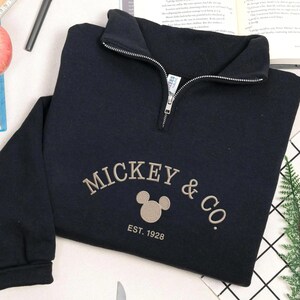 K&ouml;nnte beinhalten: Ein marineblaues Sweatshirt mit einem Viertel-Rei&szlig;verschluss am Ausschnitt. Auf der Vorderseite befindet sich der gestickte Schriftzug "MICKEY & CO." mit einer Mickey Mouse-Kopf-Grafik und "EST. 1928" darunter. Das Sweatshirt ist auf einer wei&szlig;en Oberfl&auml;che gefaltet.