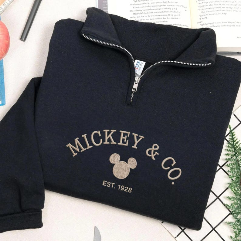 K&ouml;nnte beinhalten: Ein marineblaues Sweatshirt mit einem Viertel-Rei&szlig;verschluss am Ausschnitt. Auf der Vorderseite befindet sich der gestickte Schriftzug "MICKEY & CO." mit einer Mickey Mouse-Kopf-Grafik und "EST. 1928" darunter. Das Sweatshirt ist auf einer wei&szlig;en Oberfl&auml;che gefaltet.