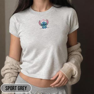 Puede incluir: Camiseta gris deportiva con un peque&ntilde;o dise&ntilde;o bordado de Stitch. El dise&ntilde;o presenta al personaje alien&iacute;gena azul con corazones rosas sobre su cabeza. La camiseta tiene mangas cortas y cuello redondo. El texto "SPORT GREY" est&aacute; en la parte inferior de la imagen.