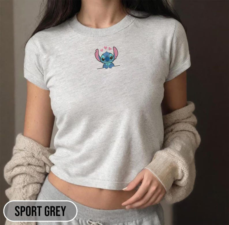 Puede incluir: Camiseta gris deportiva con un peque&ntilde;o dise&ntilde;o bordado de Stitch. El dise&ntilde;o presenta al personaje alien&iacute;gena azul con corazones rosas sobre su cabeza. La camiseta tiene mangas cortas y cuello redondo. El texto "SPORT GREY" est&aacute; en la parte inferior de la imagen.