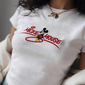 Op de afbeelding: Wit T-shirt met een geborduurd Mickey Mouse-ontwerp. Het ontwerp bevat de woorden "Mickey Mouse" in rode letters en een cartoonafbeelding van Mickey Mouse. Het shirt heeft korte mouwen.