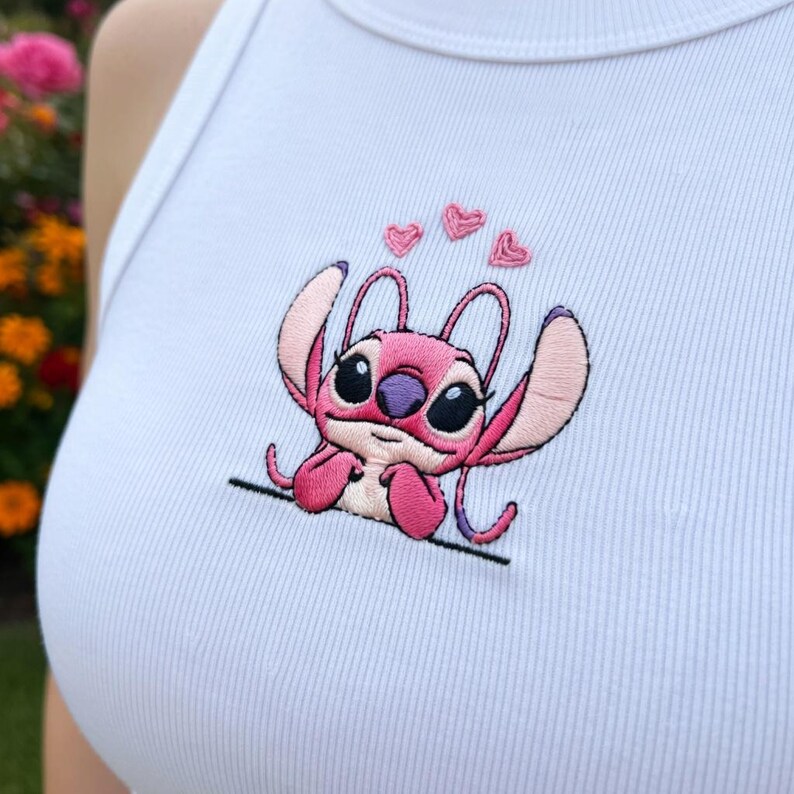 May include: White ribbed vest top with an embroidered pink cartoon character, large black eyes, and three pink hearts. The character is seated with outstretched arms. The design is on the front.