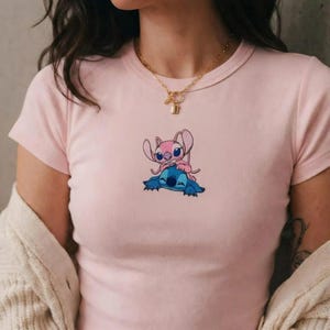 May include: A pale pink t-shirt with a cartoon illustration of two characters, one pink and one blue. The pink character is positioned on top of the blue character. The top has a round neck and short sleeves.