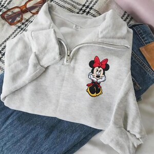 以下が含まれることがあります： ライトグレーのスウェットシャツ。ハーフジップのネックラインとミニーマウスの刺繍デザインが施されています。漫画のキャラクターは赤いリボン、赤いドレス、黄色の靴を身に着けています。スウェットシャツはブルージーンズの上に折りたたまれています。