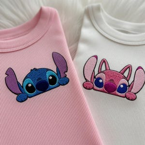 Peut inclure: Deux hauts en tricot c&ocirc;tel&eacute;, un rose et un blanc, chacun avec un personnage de dessin anim&eacute; brod&eacute;. Le haut rose a un personnage bleu, tandis que le haut blanc a un personnage rose. Les deux personnages ont de grands yeux et des oreilles.