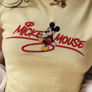 Op de afbeelding: Een lichtgeel T-shirt met een geborduurd Mickey Mouse-ontwerp. Het stripfiguur is afgebeeld in een klassieke pose, met rode en gele details. De woorden "Mickey Mouse" zijn geborduurd in rood.
