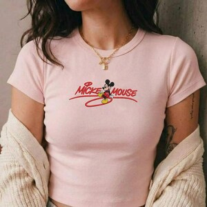 Op de afbeelding: Een lichtroze t-shirt met de tekst "Mickey Mouse" in rode letters en een cartoonafbeelding van Mickey Mouse. Het shirt heeft korte mouwen en een ronde hals.