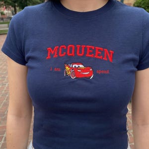 May include: Navy blue crop top featuring the name "MCQUEEN" in red embroidered lettering. Below the name is a red cartoon car graphic and the words "i am spexd". The shirt has short sleeves.