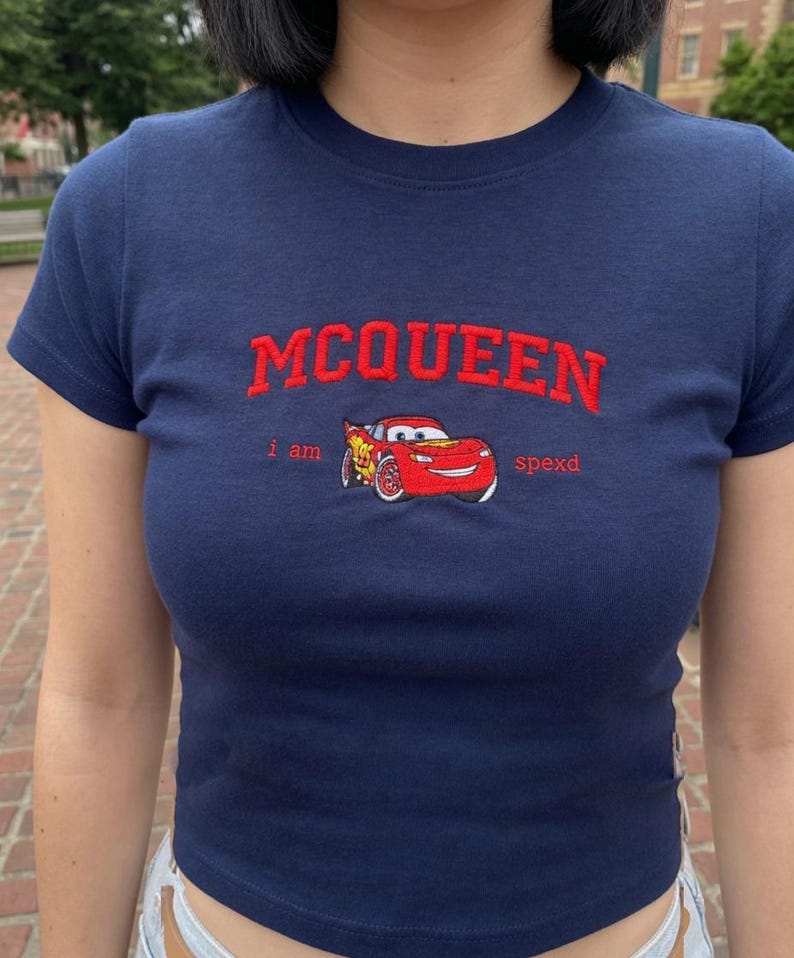 May include: Navy blue crop top featuring the name "MCQUEEN" in red embroidered lettering. Below the name is a red cartoon car graphic and the words "i am spexd". The shirt has short sleeves.