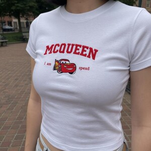 May include: White, short-sleeved t-shirt featuring the word "MCQUEEN" in red arched lettering. Below the text is a graphic of a red race car with the words "i am spexd". The shirt is made of a soft material.