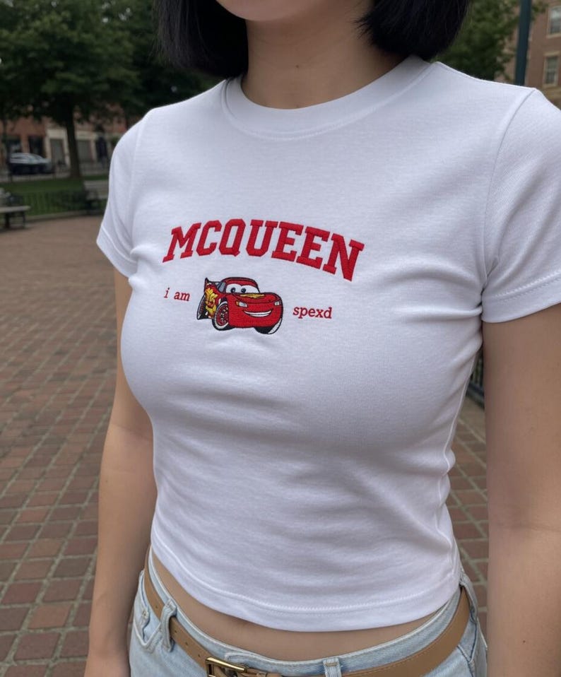 May include: White, short-sleeved t-shirt featuring the word "MCQUEEN" in red arched lettering. Below the text is a graphic of a red race car with the words "i am spexd". The shirt is made of a soft material.