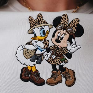 Pu&ograve; includere: Design ricamato con Paperino e Minnie Mouse in tenute safari. Paperino indossa un cappello safari, un gilet e stivali. Minnie Mouse indossa un vestito con stampa leopardata, occhiali e stivali. Il design &egrave; su sfondo bianco.